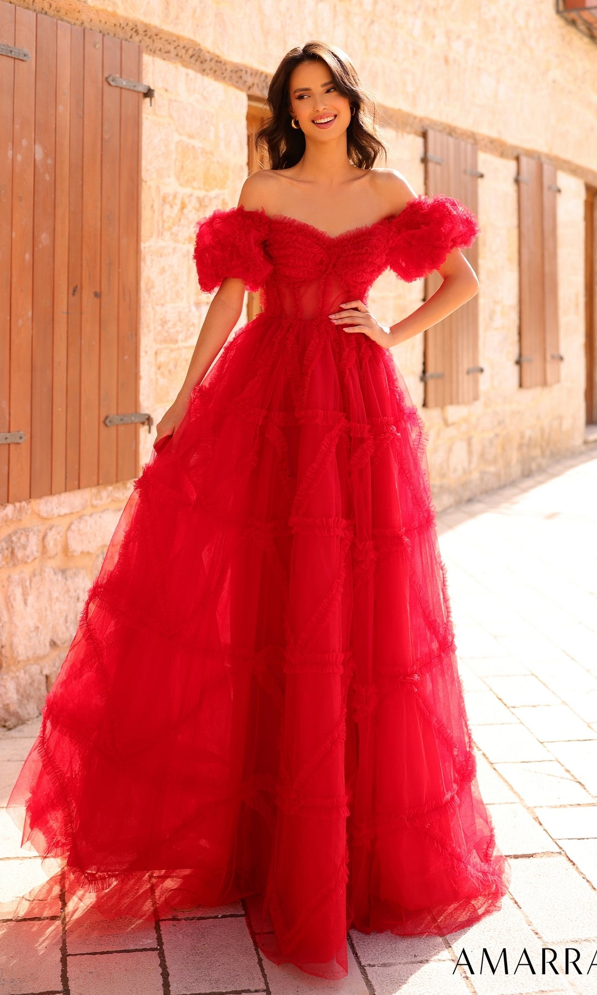 A woman stands outdoors in the sun, smiling and posing on a stone walkway in the Amarra Puff-Sleeve Long Ruffled Prom Dress 94002—an elegant, off-shoulder, floor-length red gown with tulle ruffles and dramatic puff sleeves.